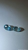 Pair of oval cut natural Cambodian zircon gemstones in seafoam blue color on a light surface, shown with clear faceting and VS clarity.
