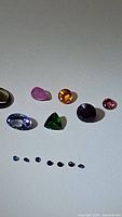Top-down photo showing a collection of 12 assorted cut natural gemstones including green, purple-blue, pink, orange-yellow, golden-brown, and blue sapphires.