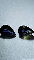 Photo showing the two pear cut blue sapphire gemstones side by side on a black surface under lighting to highlight their color and facets.
