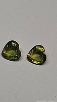 Photo showing two heart-shaped chartreuse natural sapphires on a grey background, highlighting their color and cut.