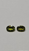 Two natural vivid apple green oval cut peridot gemstones placed side by side on neutral background.