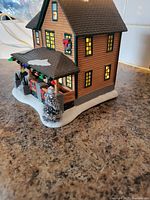 Side view of lighted Ralphie's house figurine showing yellow-lit windows, snow-topped roof, front porch with colorful Christmas lights and frosted pine tree.