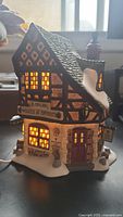 Front view of illuminated Department 56 Dickens' Village house showing detailed Tudor-style facade, signage, chimney, window lighting and snow accents.