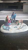 Miniature ice fishing figurine scene with two adults, child, dog, fishing hole and accessories on snow base.