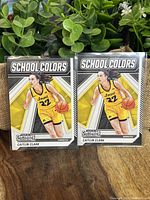 Two Caitlin Clark School Colors rookie basketball cards in clear sleeves on a wooden surface with a green plant background.