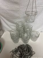 Stack of multiple clear glass candle holders with frosted designs and a pile of silver metal hanging chains in front.