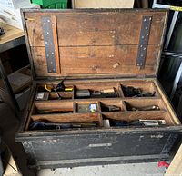Open antique wooden tool chest showing the upper compartment with various hand tools including chisels, pliers, spirit levels, and other smaller tools organized in divided sections.