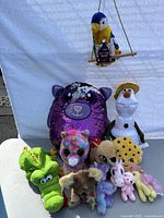 Full lot of assorted stuffed toys arranged together showing a variety of characters including a purple sequined animal, green alligator puppet, small unicorn, snowman with hat, plush parrot among others.