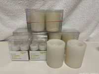 Eight white wax pillar candles grouped with four boxes of LED battery operated tea light candles, two pillar candles outside packaging.
