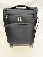Front view showing IT logo, two zippered compartments, and four spinner wheels.