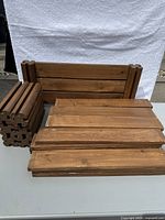 Photo showing stained cedar garden bed boards stacked flat and six corner end pieces stacked vertically. Boards have a rough surface with some nail holes and staining.