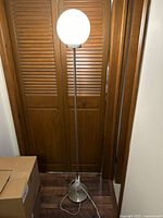 Full view of floor lamp standing on wood floor against wooden panels, showing metal base, long metal pole and white frosted globe light diffuser at the top