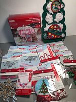 Photo showing 3D candy shop craft kit box, paintable plaster Christmas ornaments on a green tree-shaped holder, assorted cross-stitch kits, candy cane shaped craft pieces, and embellished patch stickers arranged on a surface.
