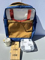 Photo showing all four new IKEA items: mustard yellow and blue backpack, white candle in glass jar, BESTÄMMA glass food container with label, and wooden phone holder.