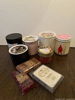 Photo of six candles in jars and one tin along with three packs of wax melts in front of a white wall on a wooden surface