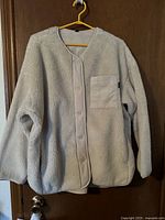 Light grey fleece jacket shown hanging on a hanger, buttoned front, collarless, long sleeves, with light beige fabric patch pocket.