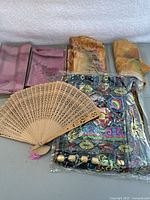 Photo showing 5 folded hand-painted silk scarves behind an embroidered bag and a wooden carved fan.