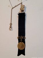 Full view of the antique pocket watch fob with black ribbon, gold-tone clip, chain, and medallion pendant lying flat showing entire length