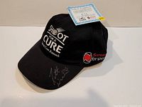 Black baseball cap showing 'Shot For A Cure' embroidery, 'Sound For Sport' logo, player autograph on brim, and certificate of authenticity laid behind.