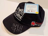 Black baseball cap with charity logos and Certificate of Authenticity visible on top of the cap. Autograph in silver on brim.
