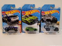 Three Hot Wheels cars in original packaging: a black Chevy Silverado Trail Boss, a lime green Ford Focus RS, and a grey Jeep Wrangler.