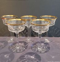 Five antique stemmed glasses with clear etched bowls and gold gilt rims arranged in a group.