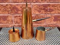 Front angle of the tall copper coffee pot, creamer jug, and sugar bowl with wooden handles and pointed lid on a patterned surface against a brick wall.