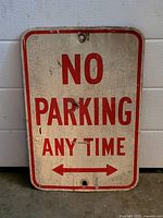 Photo of a rectangular vintage metal sign. The sign has a white background with a red border. Red text reads 'NO PARKING ANY TIME' with a red double-headed arrow at the bottom. Surface shows significant wear, scratches, and cracking indicating age and use. Two mounting holes visible at top and bottom center.