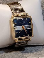 Square vintage wristwatch with a black dial featuring a white horizontal stripe, gold-tone case and mesh band, showing date and time.