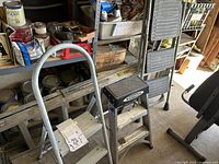 All four ladders arranged in garage setting with paint cans and other tools in background. One 8-foot aluminum extension ladder and three stepladders of varying styles and sizes visible.