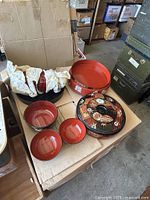 Seven Asian acrylic dishes arranged on a cardboard box showing red lacquered bowls and platters, and a black lacquered dish with decorated lid.