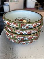 Stack of three round ceramic plates with detailed fruit and floral Asian designs, displaying red grapes and green vines inside oval panels on a white background and bordered with green and gold trim.