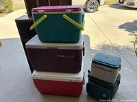 Photo showing stacked coolers: purple Coleman cooler, white/red cooler, and green Igloo cooler with yellow handles and burgundy lid on top. Two smaller coolers (white/blue Coleman and small dark green) adjacent.