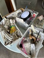 Photo showing baskets containing white extension cords, small hardware items, packaged sink strainers, and a round light fixture cover on a table.