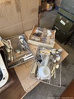 Two wire drawer trays with assorted used flatware including forks, knives, spoons, and serving utensils, displayed on cardboard boxes in a storage area.