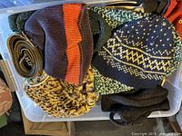 Plastic bin filled with assorted knit winter hats in multiple colors and patterns including striped and geometric designs, along with knit accessories and scarves.