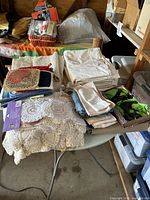 Photo showing folded assorted linens including white cloths, crocheted doilies, folded napkins, and a box with potholders and napkin holders.