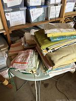 Photo of assorted linens stacked on a small circular table in a storage area, showing sheets, aprons, and kitchen towels.