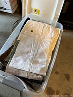 Multiple men's dress pants folded in a plastic bin, showing neutral colors like cream and brown.