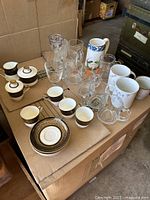 Overview of entire lot showing tea cups, saucers, assorted clear glass mugs and drinking glasses, ceramic coffee cups, and a ceramic pitcher with a floral and fruit pattern.