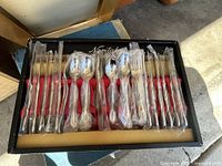 Set of stainless steel flatware pieces individually wrapped in plastic, arranged neatly in Noritake box with red interior.