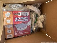 Cardboard box containing plastic utensils, paper plates, and microwave cooking covers packed in plastic bags and original packaging. Contents are assorted disposable dining items.