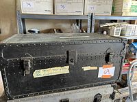 Front view of black metal foot locker showing riveted edges, two latches, central lock, and handwritten paper label taped on front.