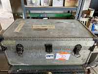 Front view of the foot locker trunk showing metal riveted construction, three metal latches (center missing loop), stickers and signs of wear.