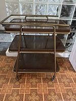 Front view of vintage 3-tier bar cart showing brass-tone metal frame, three wood-look shelves, and caster wheels.