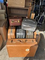Nine vintage suitcases stacked showing various colors and styles including hard cases and train cases, placed in a garage or storage area.
