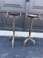 Pair of two heavy wrought iron candle holders with three-legged bases and ornate hook details on top, shown side by side on asphalt surface.