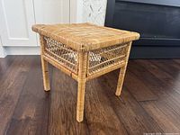Front angle showing square rattan side table with woven top and open diamond pattern sides, against a hardwood floor and near a fireplace.