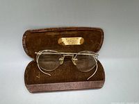 Pair of antique gold framed bifocal eyeglasses displayed open inside brown velvet lined leather case showing interior label.