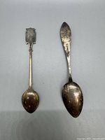 Photo of two silver spoons on gray background, one with a decorative enamel crest handle, the other with floral engravings